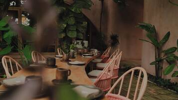 A dining area with a long wooden table, wicker chairs, and potted plants.