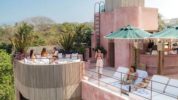 Un área de lounge en la azotea con sillas blancas y sombrillas verdes, rodeada de abundante vegetación y un edificio rosa.