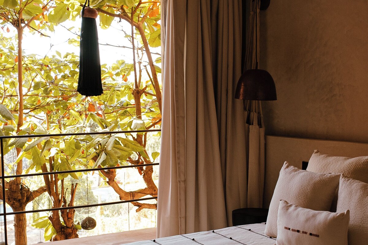 Un dormitorio con cama, una lámpara colgante, vistas a árboles y una ventana con cortinas.
