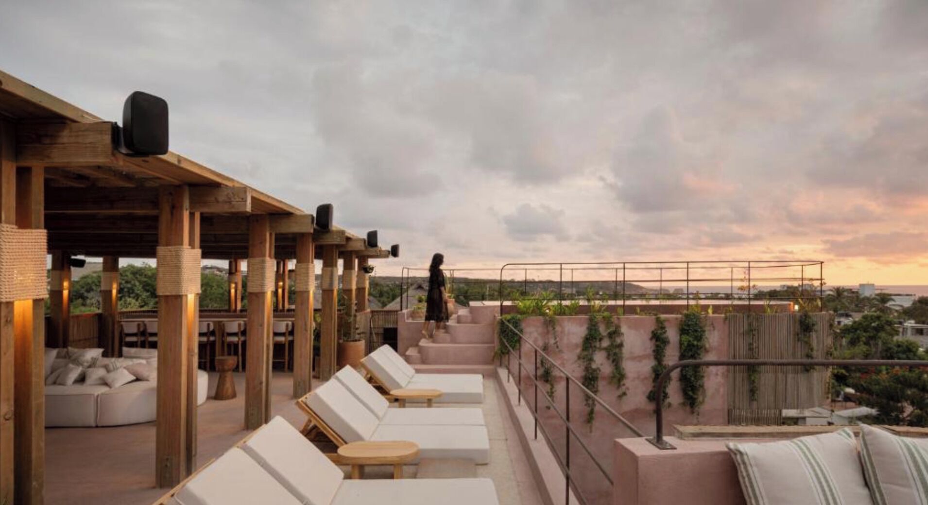 Un salón en la azotea con sillones blancos, pérgolas de madera y vistas a la ciudad al atardecer.