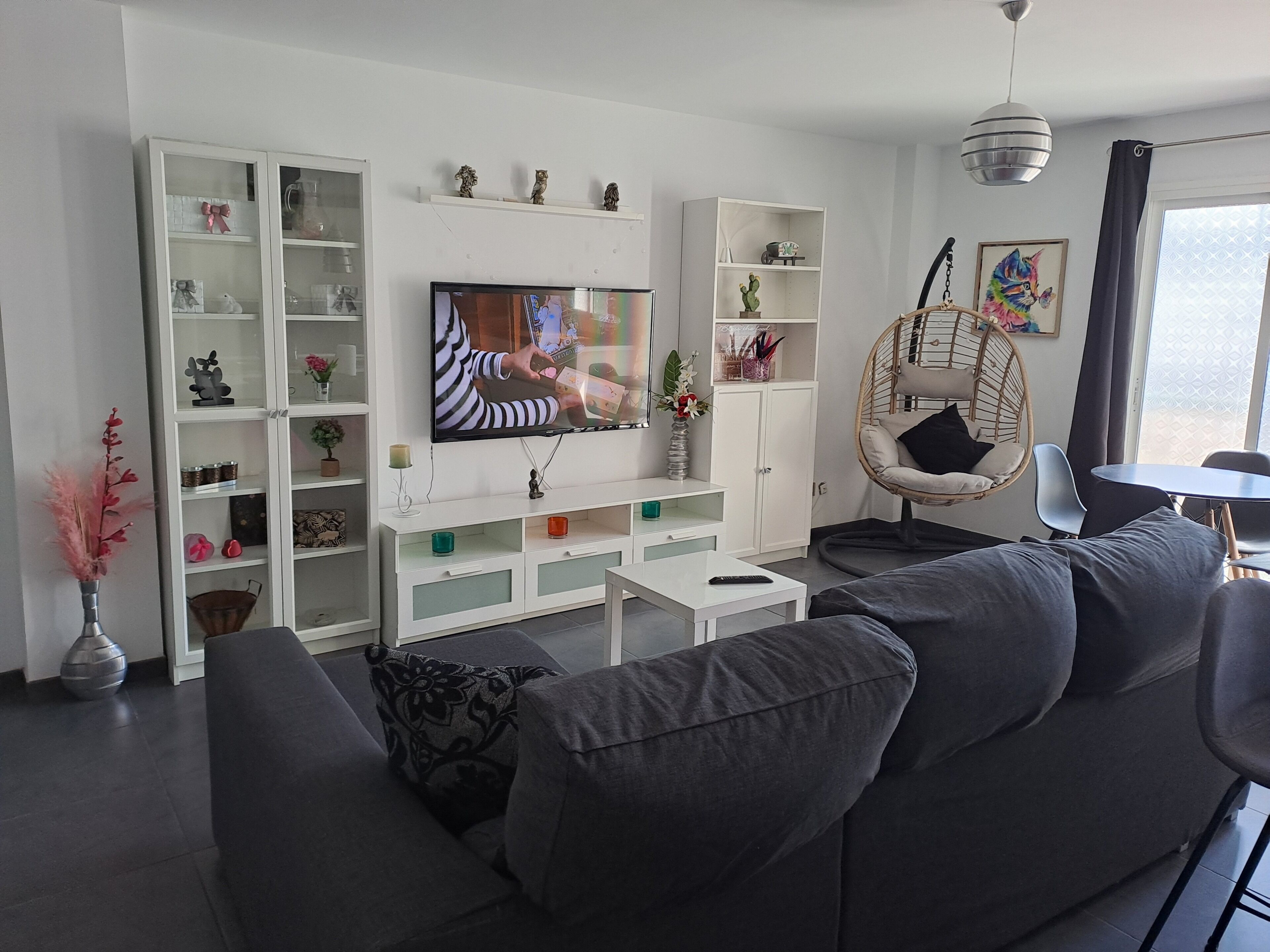 Een moderne woonkamer met een grote flatscreen televisie, een witte tv-meubel, een grijze bank, een hangstoel en een eethoek met een tafel en stoelen.