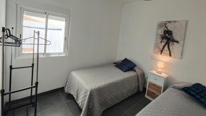 Una habitación con dos camas, una ventana con cortinas, un cuadro colgado en la pared y una mesita de noche con lámpara.