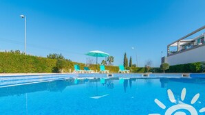 Ein klares, blaues Schwimmbecken mit Liegestühlen und Sonnenschirm, umgeben von Grünflächen und modernen Gebäuden.