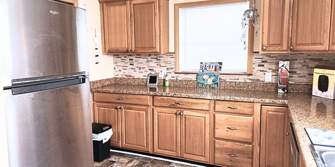 A kitchen with wooden cabinets, a stainless steel refrigerator, and a window with a beach-themed sign.