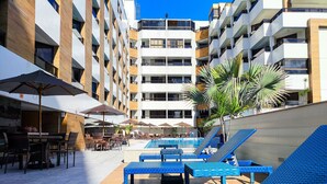 Zona de piscina con sillones de descanso y sombrillas, una piscina y un edificio con varios balcones.