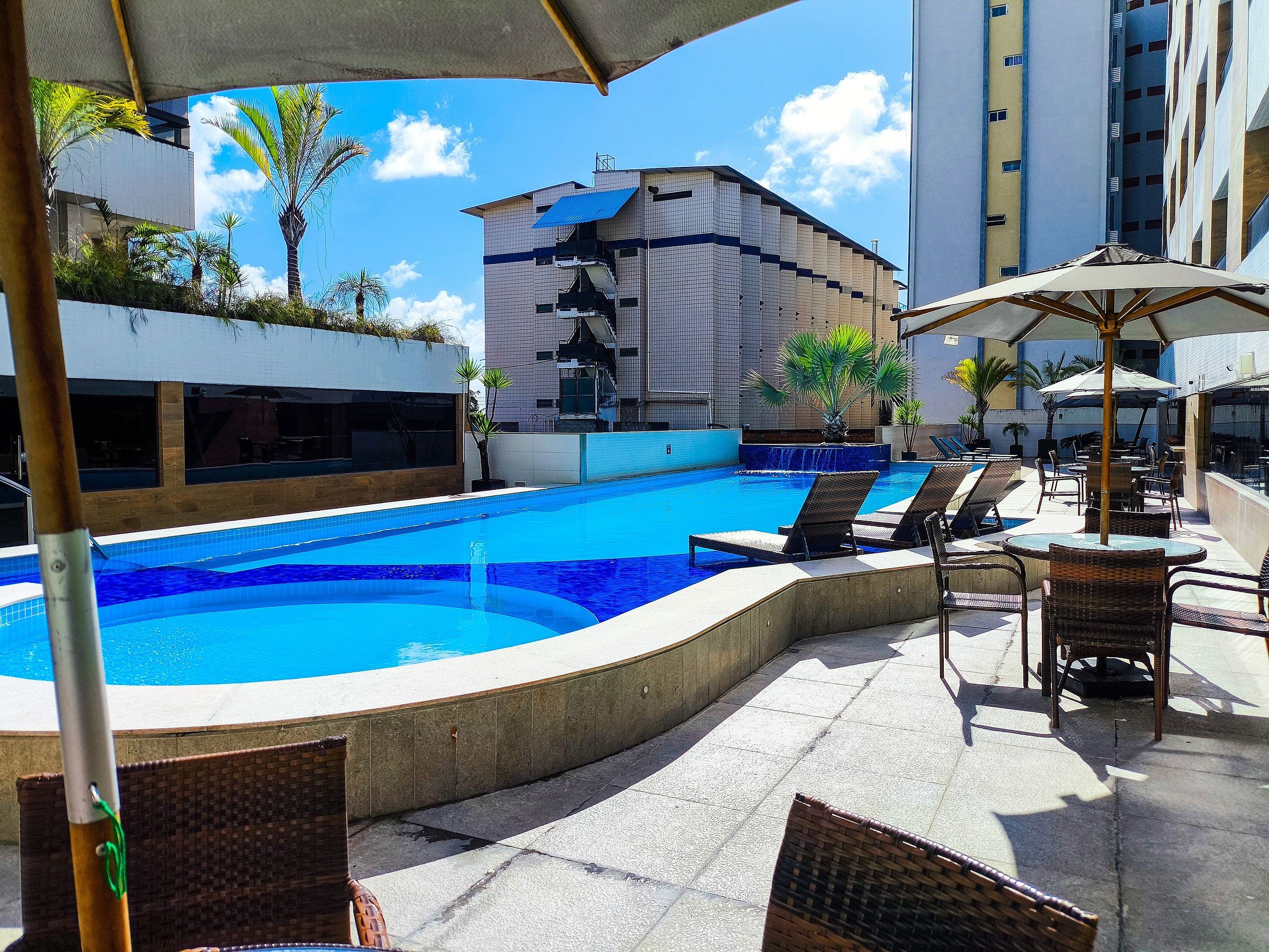 Piscina con sillones de descanso y sombrillas, rodeada de edificios y palmeras.