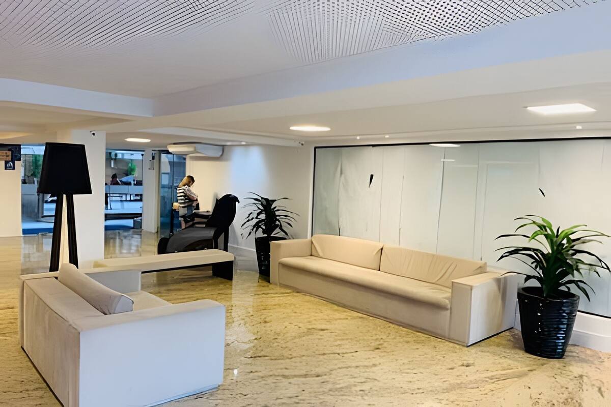A modern lobby with a beige sofa, a white armchair, and a floor lamp. There are two potted plants and a large window with a view of the outside.
