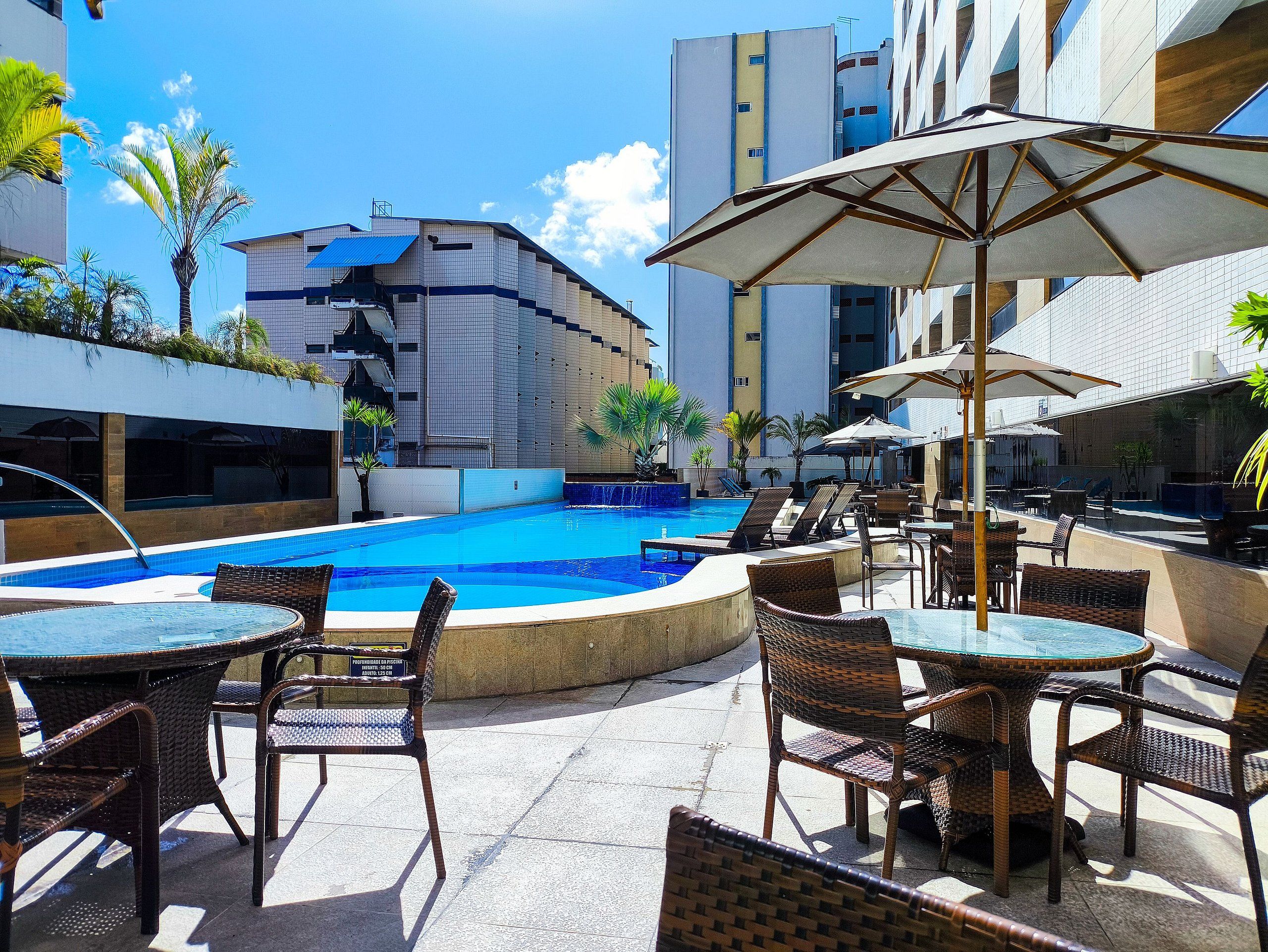 Área de piscina no terraço com espreguiçadeiras e guarda-sóis, cercada por edifícios modernos.