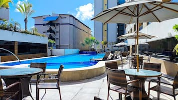 Área de piscina no terraço com espreguiçadeiras e guarda-sóis, cercada por edifícios modernos.