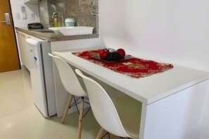 Una pequeña cocina con una encimera blanca, dos sillas blancas y un mantel individual rojo con frutas.