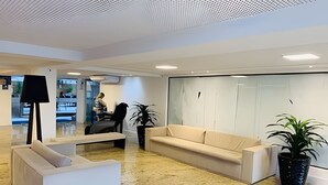 Un vestíbulo moderno con un sofá color beige, un sillón blanco y una lámpara de pie. Se ven dos plantas en macetas y un amplio ventanal con vistas al exterior.