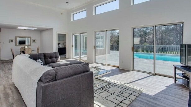A spacious living room with a grey sectional sofa, a flat-screen TV, and a large sliding glass door leading to a pool. - Hamptons getaway (East Quogue)