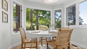 A dining area with a round table and four wicker chairs, large windows with a view of trees, and a white ceiling light.