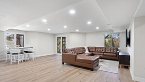 A spacious living room with a brown leather sectional sofa, a dining table with chairs, and large windows offering a view of trees.
