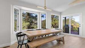 A spacious dining area with a wooden table and benches, large windows offering views of trees and mountains, and a sliding glass door leading to an outdoor deck.