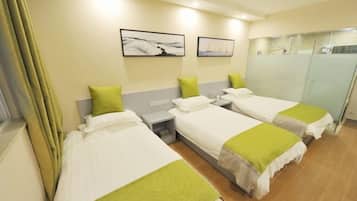 Ein Hotelzimmer mit drei Einzelbetten, jedes mit einem grünen Kissen und weißer Bettwäsche. An der Wand hängen zwei gerahmte Bilder, ein kleiner Nachttisch mit einer Lampe und ein Duschbereich mit Glastrennwand.
