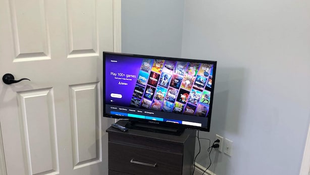A flat-screen TV on a black stand with a game library interface, a white door, and a dark wooden floor. - Shay-203-Cozy Holiday Room (Pickering)