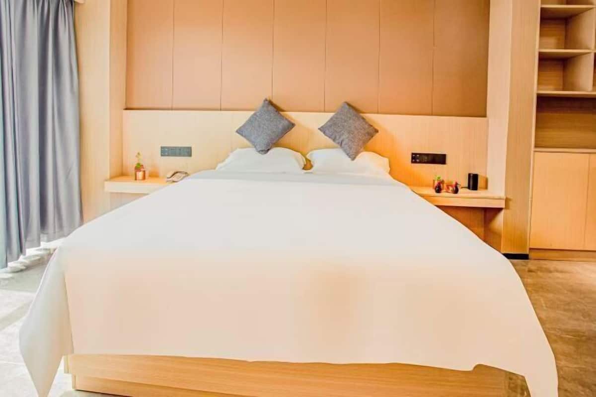 A hotel room with a large bed, a bedside table with a lamp, a small shelf with decorative items, and a wooden headboard.