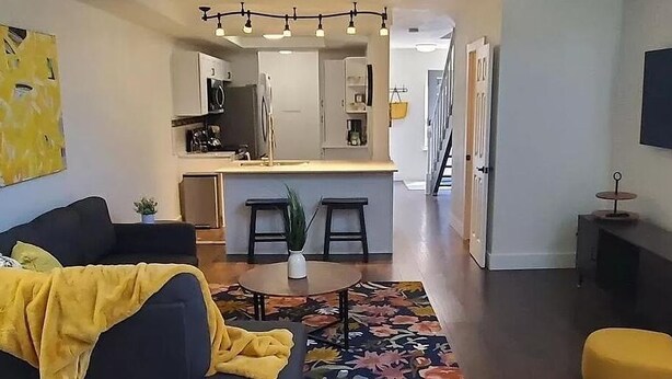 A modern living room with a sofa, a coffee table, and a yellow blanket. There is a kitchen area with a refrigerator, a microwave, and a sink. The room has a large window and a door leading to another room. - DT Boise Townhome near BSU (Boise)