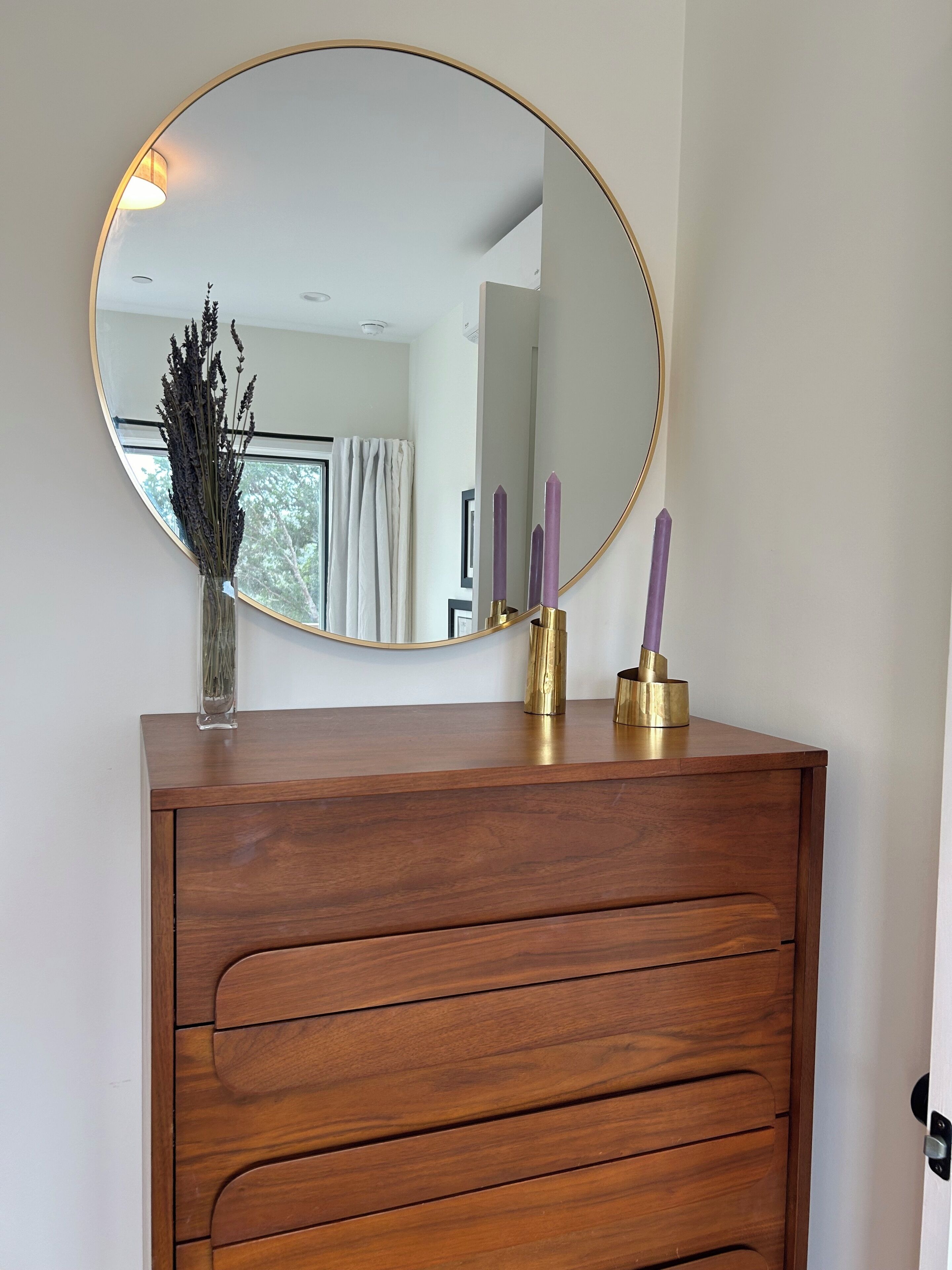 A wooden dresser with a round mirror, a vase with dried flowers, and two purple candles.