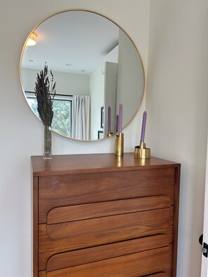 A wooden dresser with a round mirror, a vase with dried flowers, and two purple candles.