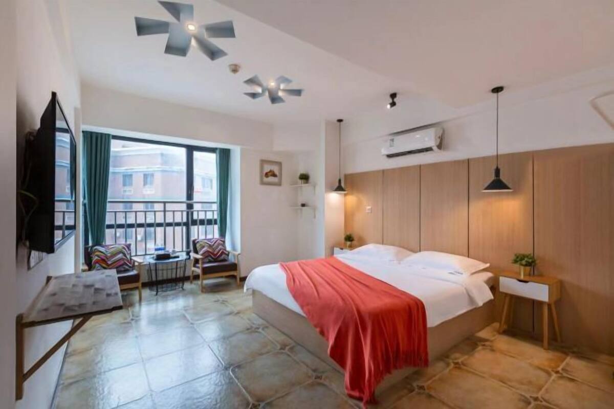 A hotel room with a bed, a small table, a chair, a TV, and a balcony with a view of buildings.