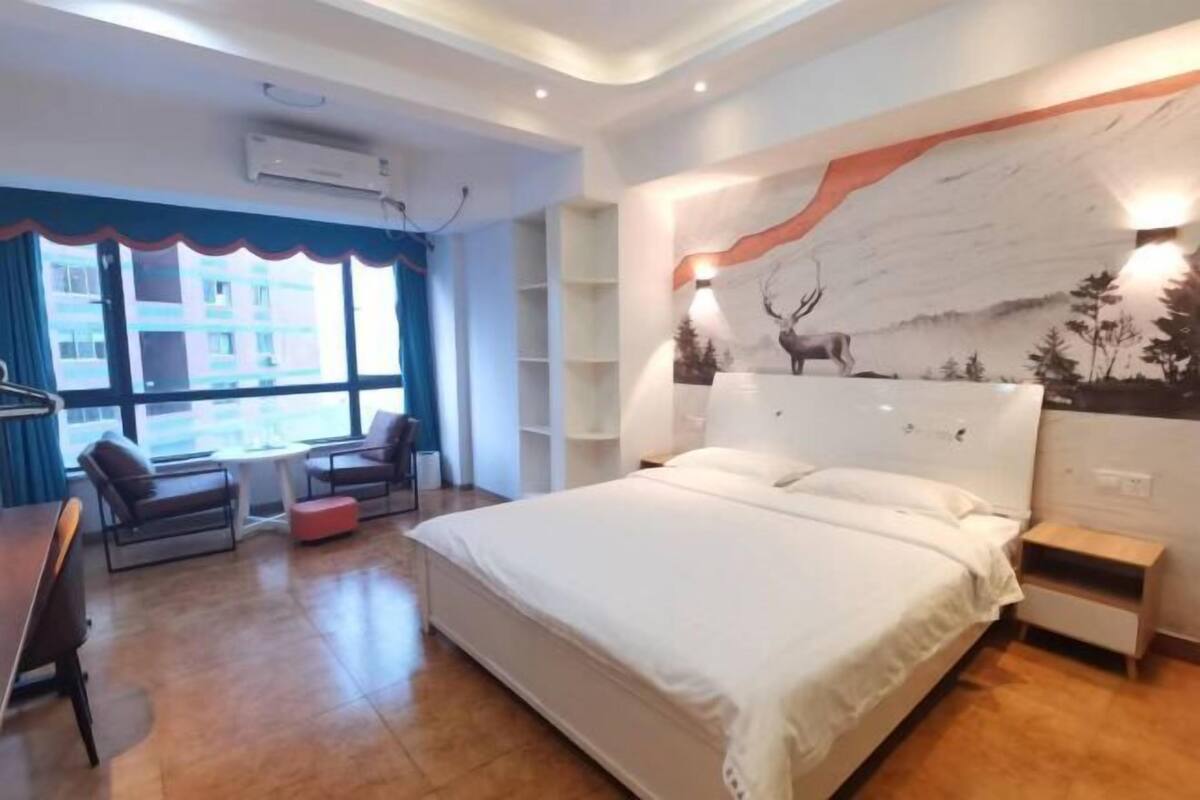 A bedroom with a large bed, a desk, and a chair. There is a window with a view of buildings outside.