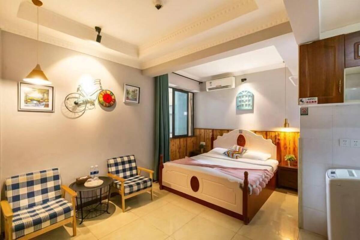 A bedroom with a bed, two chairs, a small table, a bicycle decoration, and a kitchen area.