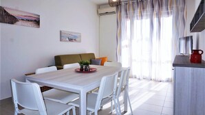 Un comedor con mesa de madera y sillas blancas, un sofá con cojines, una tetera roja sobre una encimera y vistas a través de cortinas traslúcidas.