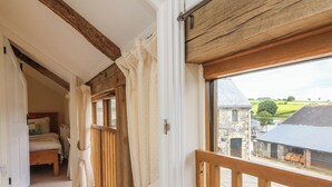 A narrow corridor with a wooden railing, leading to a bedroom with a bed and a window overlooking a rural landscape. - Hunstone Barn (South Molton)