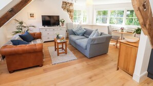 Ein gemütliches Wohnzimmer mit braunem Ledersofa, einem grauen Sofa und einem Holztisch. Ein Fernseher steht auf einer weißen Kommode, und eine Wand ist rustikal mit Stein verkleidet.