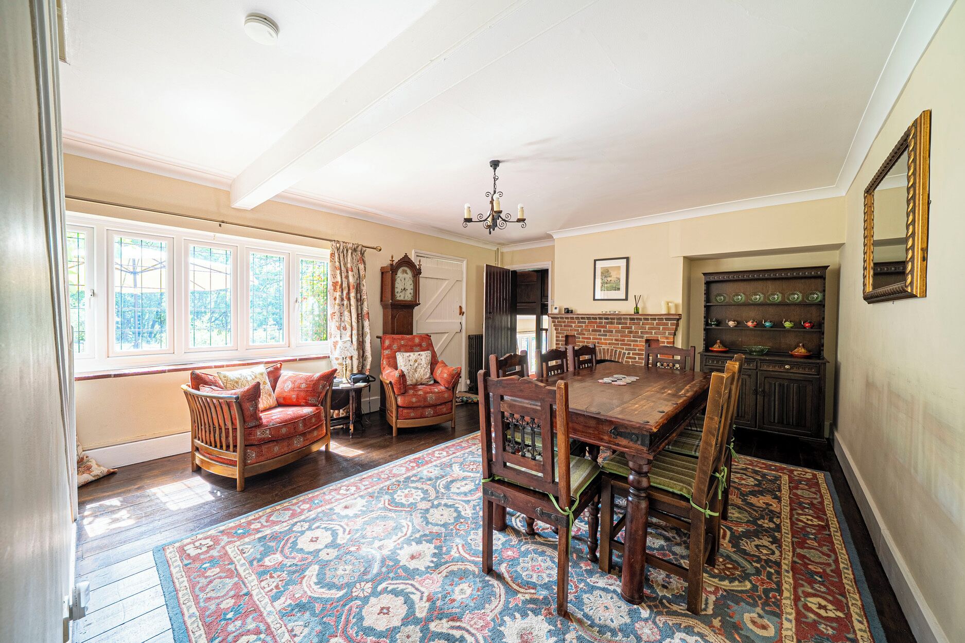 A dining room with a wooden table, chairs, a patterned rug, a clock, and a brick fireplace.
