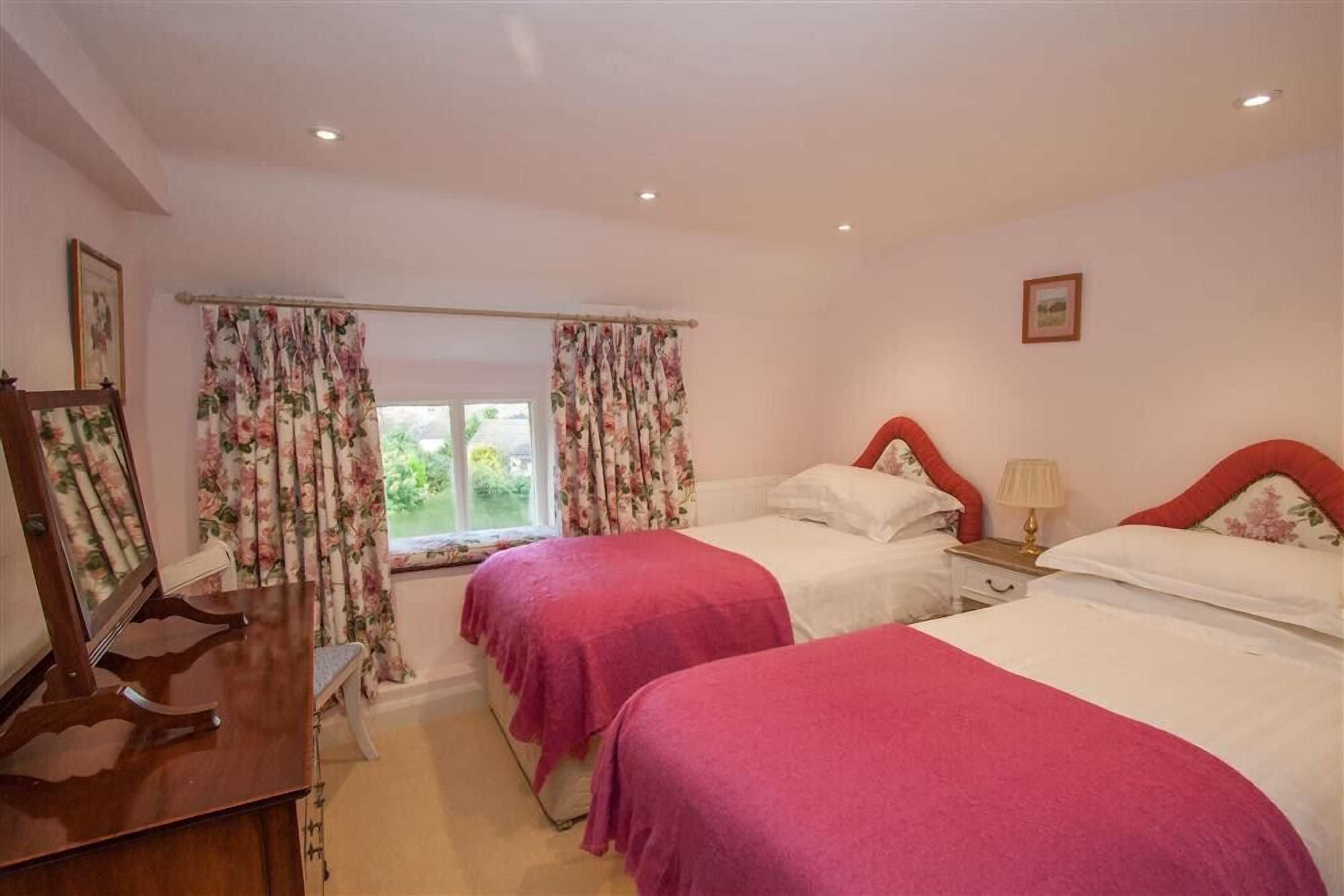 Une chambre avec deux lits, des rideaux à motifs floraux, une table de chevet avec une lampe et une commode en bois.