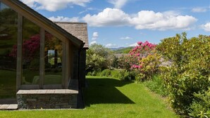 Une maison moderne dotée de grandes fenêtres en verre, d’une fondation en pierre et d’un jardin bien entretenu agrémenté de fleurs roses.