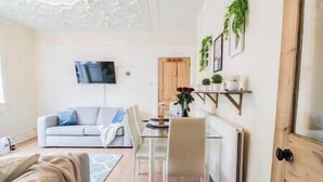 A living room with a sofa, a coffee table, and a TV mounted on the wall. There is a dining table with chairs and a radiator on the wall.