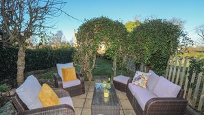 An outdoor seating area with wicker furniture and a glass-top table, surrounded by greenery and a wooden fence. - Oscar's Pad (Llanfairpwllgwyngyll)