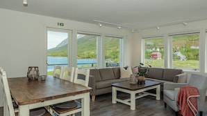 Un vaste espace de vie comprenant une table à manger en bois, des canapés et de grandes fenêtres donnant sur un lac et des montagnes.