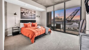 A modern hotel room with a large bed, a cityscape view, and a concrete ceiling.