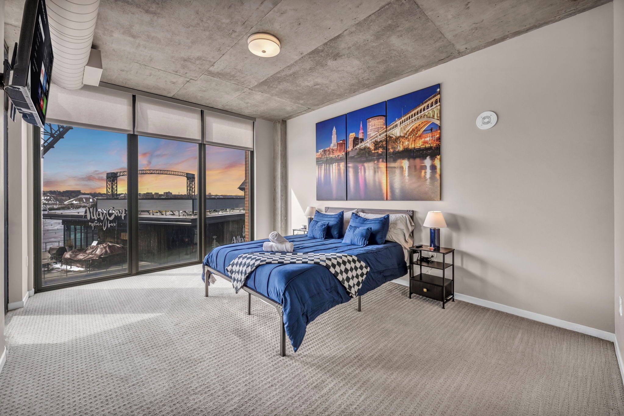 Una camera da letto con un letto grande, un comodino, una lampada e vista sulla città attraverso un'ampia finestra.