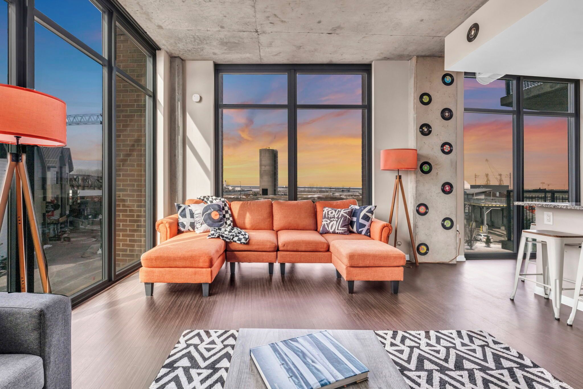 Una sala de estar moderna con un gran ventanal, sofá naranja, alfombra estampada y vistas del horizonte urbano.