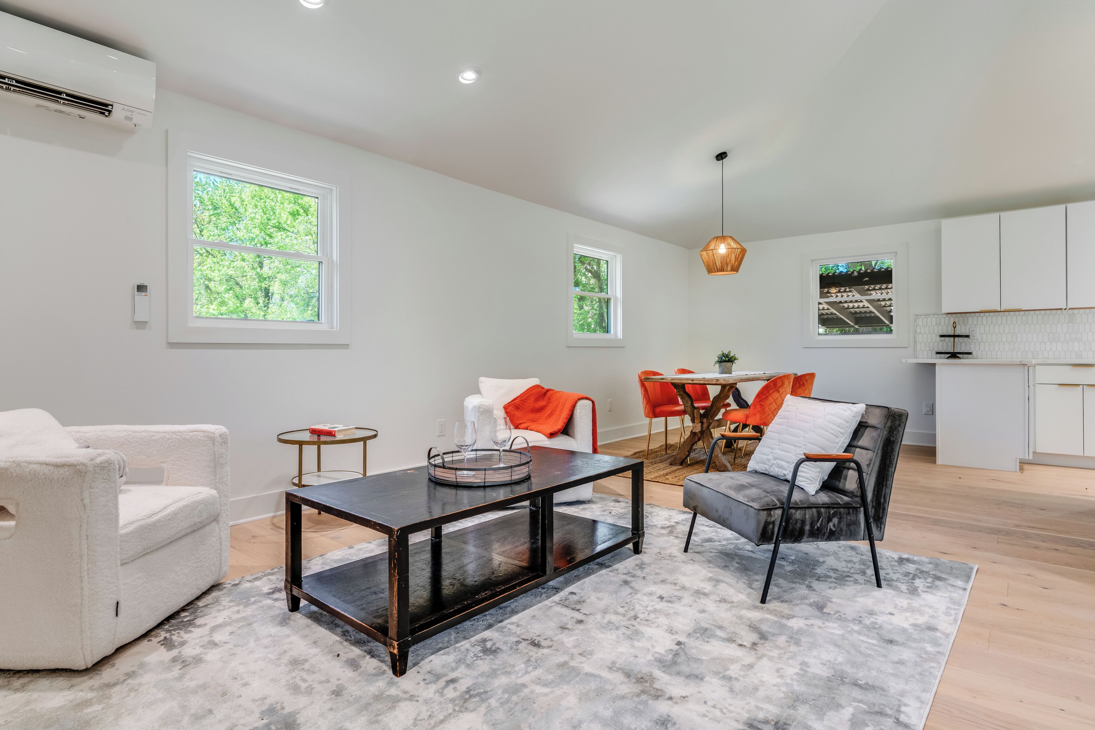 A spacious living area with a modern sofa, a coffee table, and a dining table with orange chairs. There are large windows providing natural light and a view of greenery outside.