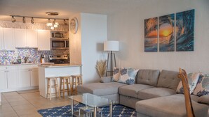 A modern kitchen with white cabinets, a microwave, and a sink. There is a dining area with stools and a glass table. The living room has a beige sofa with decorative pillows and a blue patterned rug. - Sea You Soon Retreat+Jeep option (St. Thomas)