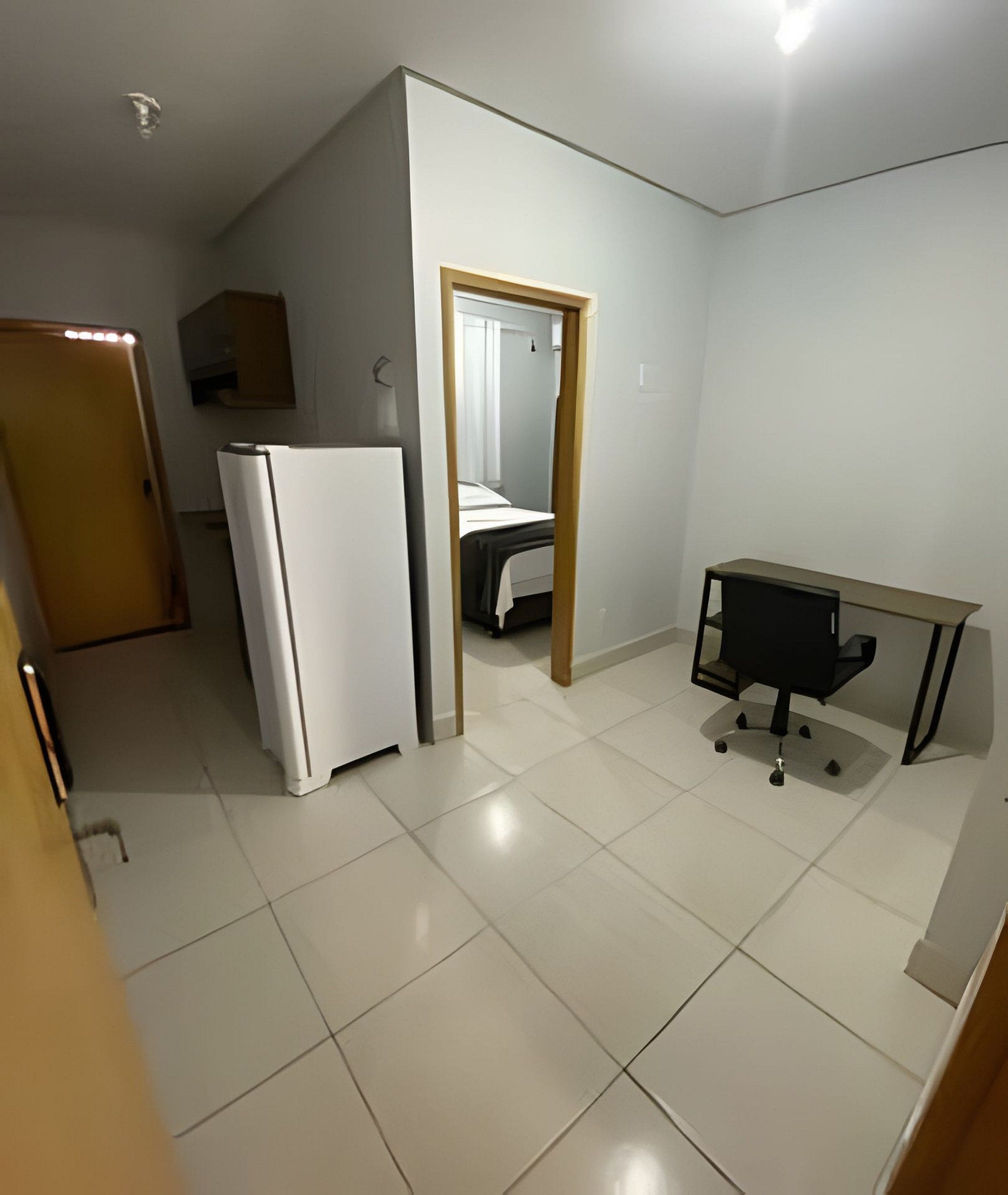 A compact room with a white refrigerator, a small desk with a chair, and a bed in the background.