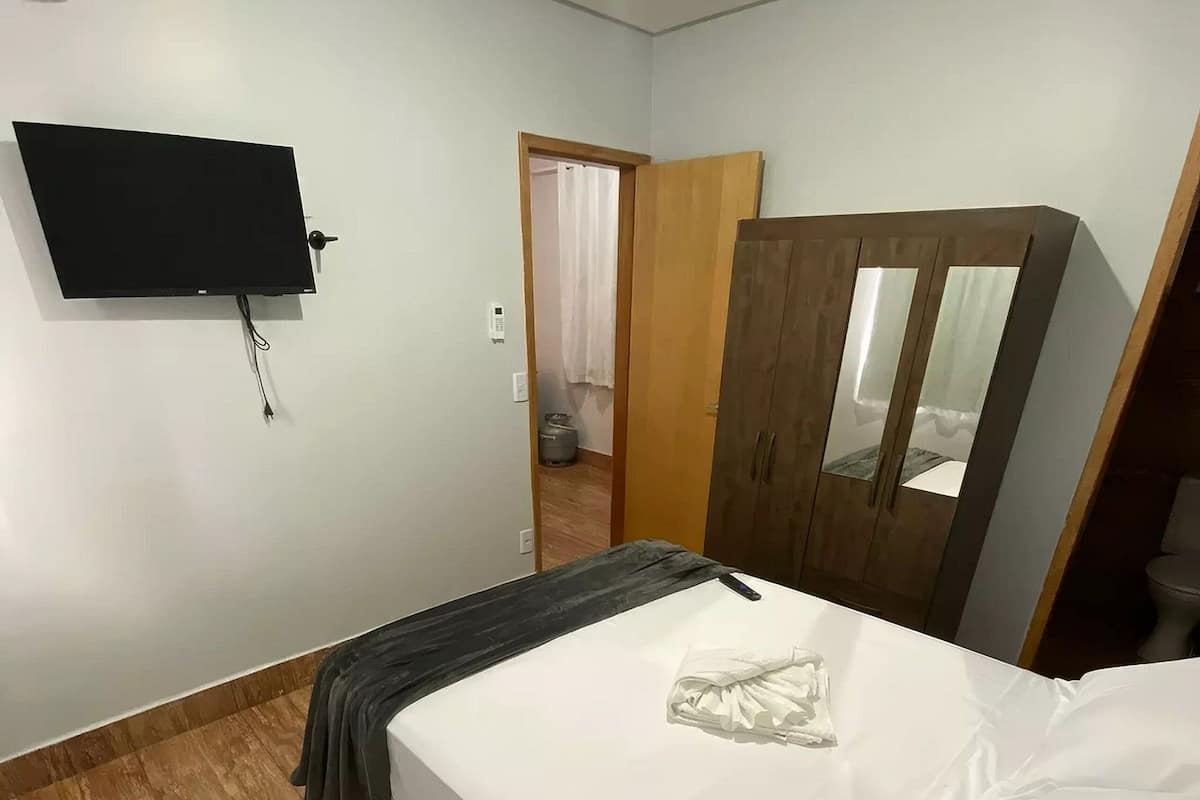 Une chambre à coucher comprenant un lit, une armoire en bois, un téléviseur fixé au mur et une porte donnant sur une autre pièce.
