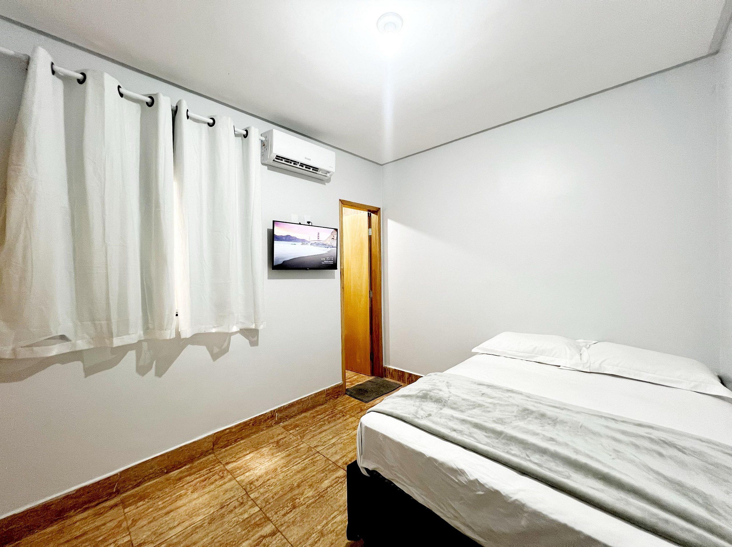 Hotellihuone, jossa on sänky, seinälle asennettu televisio ja valkoisilla verhoilla varustettu verhotanko.