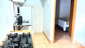 Une cuisine compacte équipée d’une cuisinière à gaz, de placards et d’un petit plan de travail. Une chambre à coucher avec un lit et une table de chevet est visible à travers une porte ouverte.