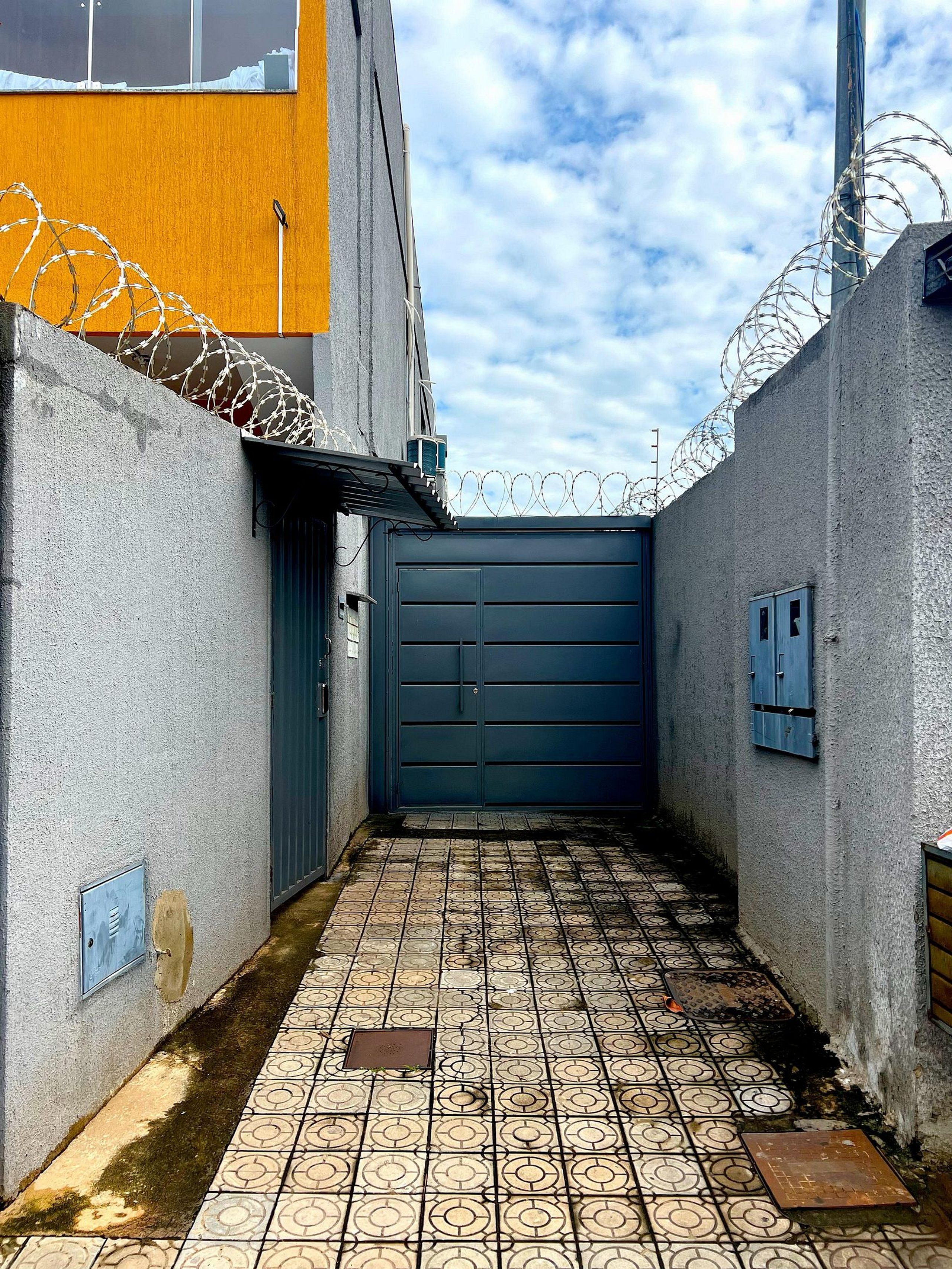 Un estrecho callejón con una puerta azul, suelo de baldosas y alambre de púas en las paredes.