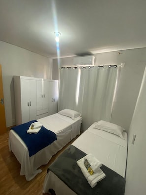 A room with two single beds, a wardrobe, a table, and a ceiling light. - EDIFICIO GERVASIO (Goiania)
