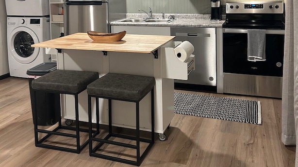 Eine kompakte Küche mit Einbauwaschmaschine, Mikrowelle, Herd und Spüle. Zwei Hocker und ein kleiner Holztisch sind vorhanden.