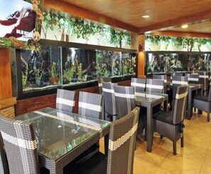 A restaurant interior with glass-top tables and wicker chairs, surrounded by large aquariums. - Safari Lodge Hotel (Cisarua)